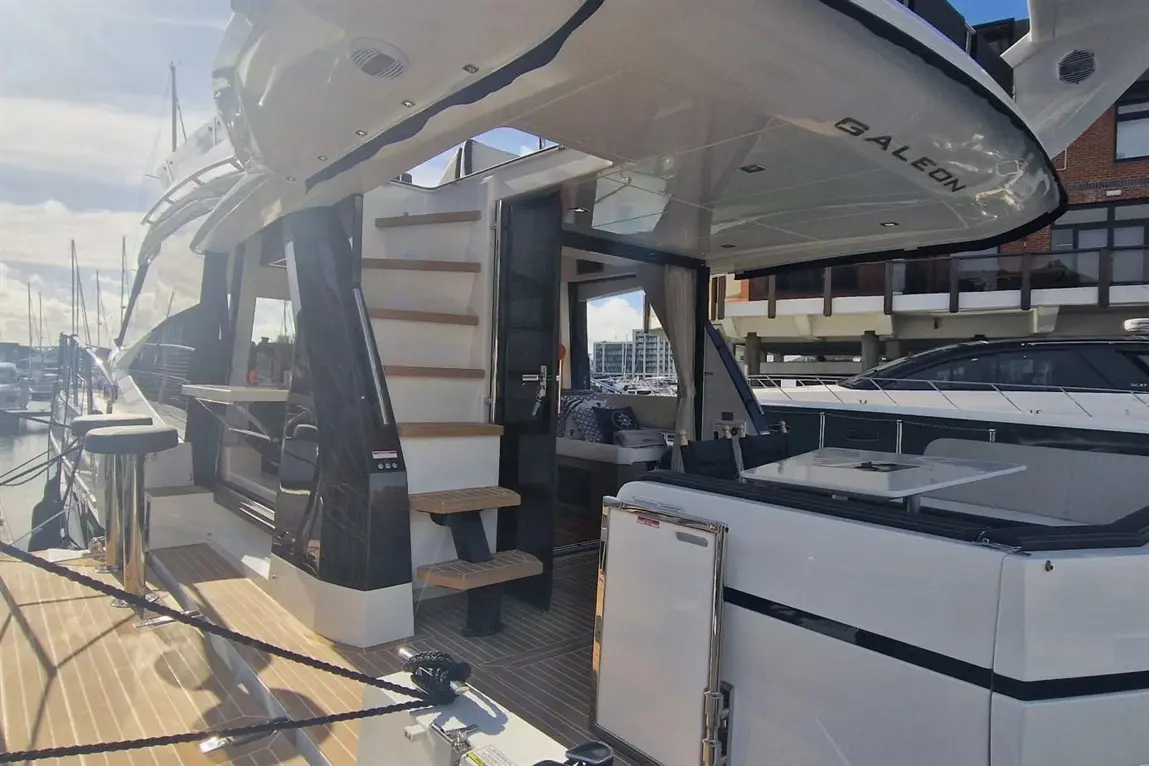 A Galleon yacht docked, showcasing a spacious deck with seating, a modern interior entrance, and boats in the marina background.